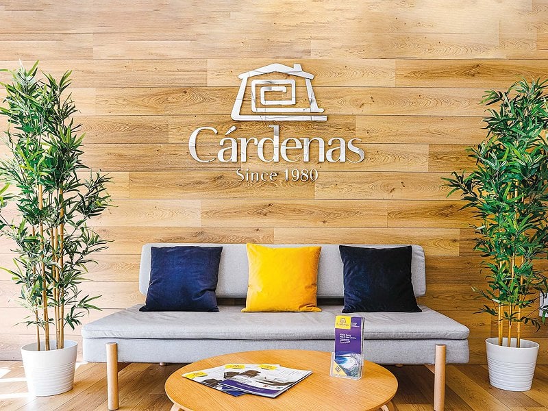 Een bank met blauwe en gele kussens en het logo van makelaar Cárdenas op een houten muur.